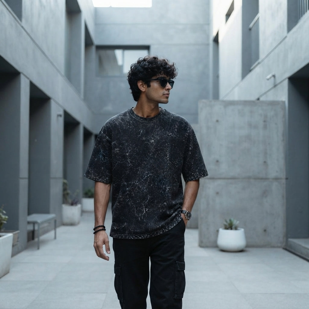 A young South Asian person wearing a charcoal acid-washed oversized t-shirt in a bright modern urban setting.