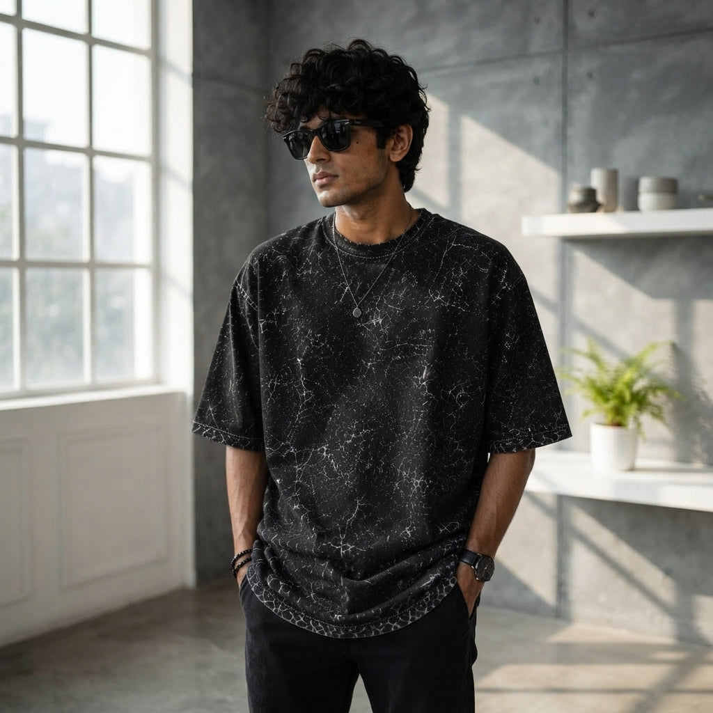 A South Asian person wearing a charcoal acid-washed oversized t-shirt in a modern, sunlit urban apartment.