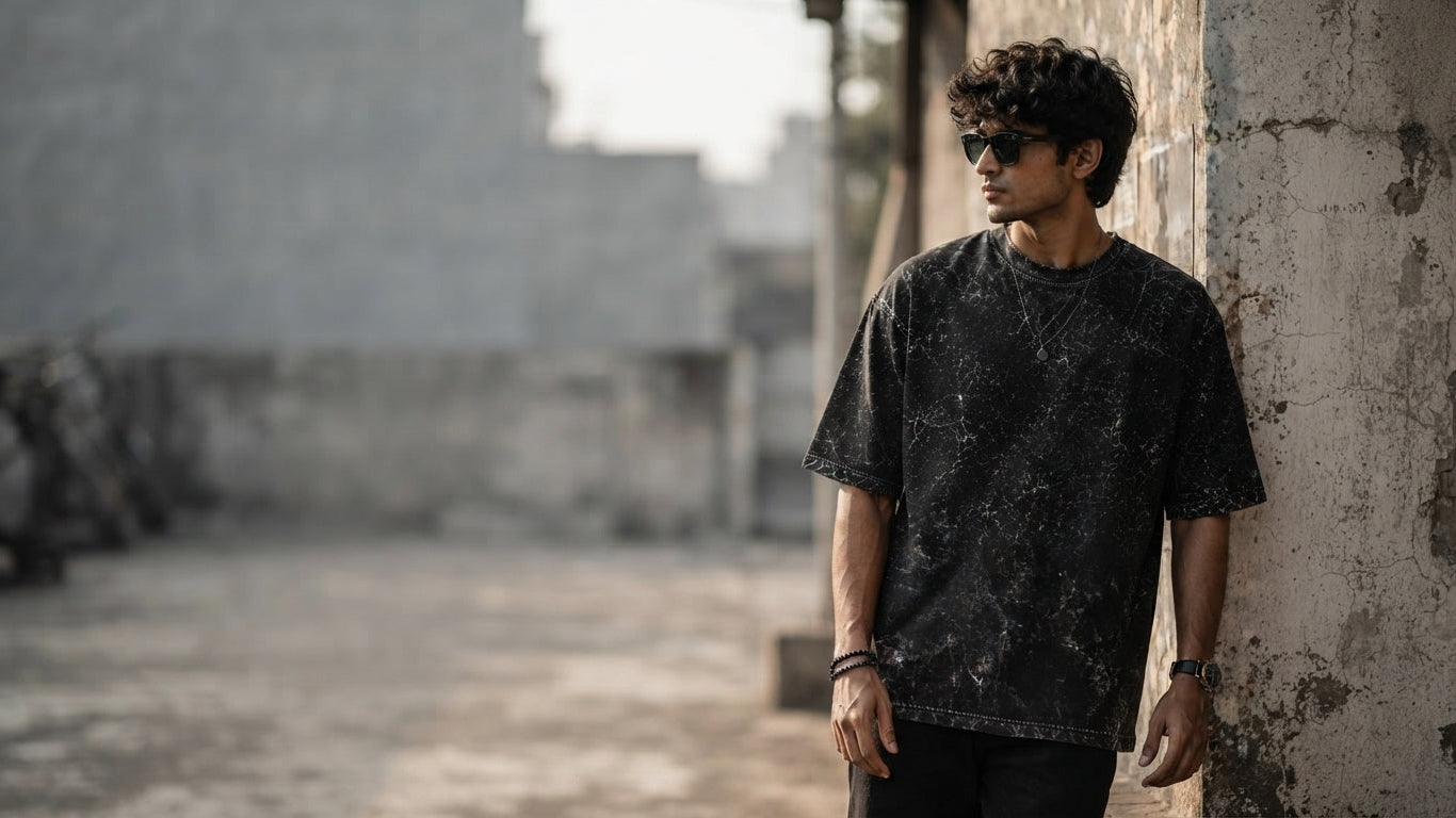 A South Asian person wearing a charcoal acid-washed oversized t-shirt in a modern urban space with soft, natural lighting.