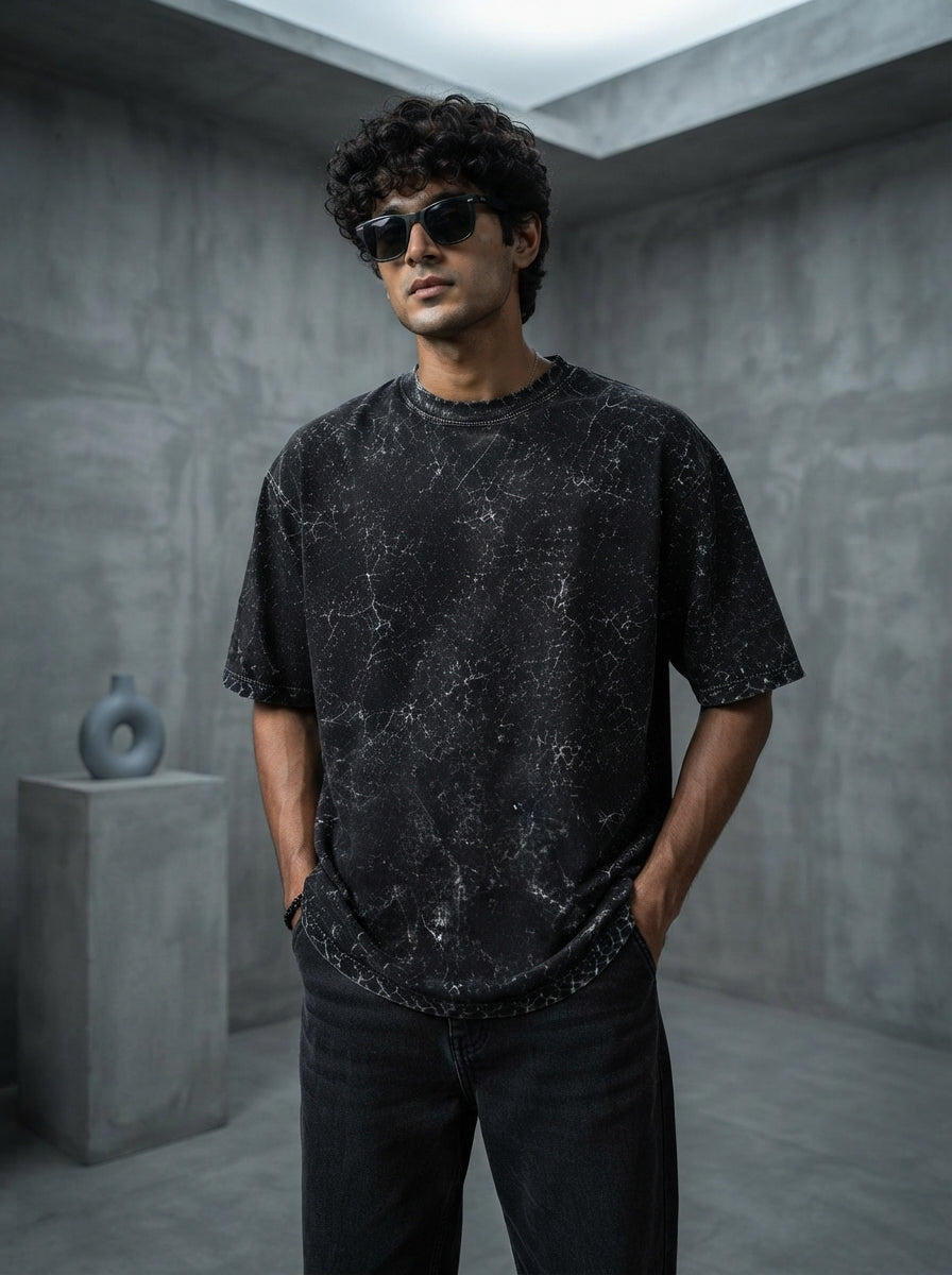 A young South Asian man wearing a charcoal heavyweight acid-washed oversized cotton t-shirt in a minimalist concrete studio.