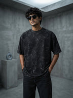 A young South Asian man wearing a charcoal heavyweight acid-washed oversized cotton t-shirt in a minimalist concrete studio.